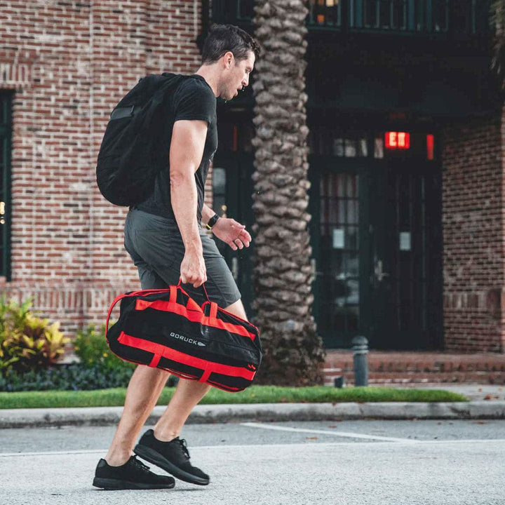 A man in a black shirt and shorts, wearing GORUCK Ballistic Trainers in Blackout + Black designed for functional fitness, walks with a backpack and carries a red duffel bag on a city sidewalk.