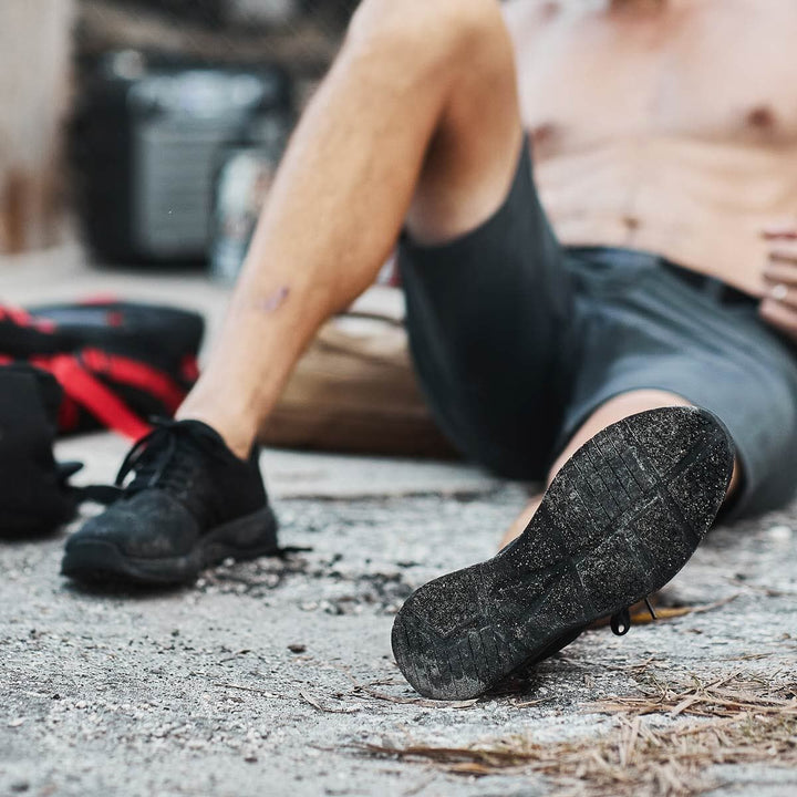 A person in shorts and sneakers sits on the ground, showcasing the soles of their Ballistic Trainers - Blackout + Black by GORUCK, perfect for functional fitness.