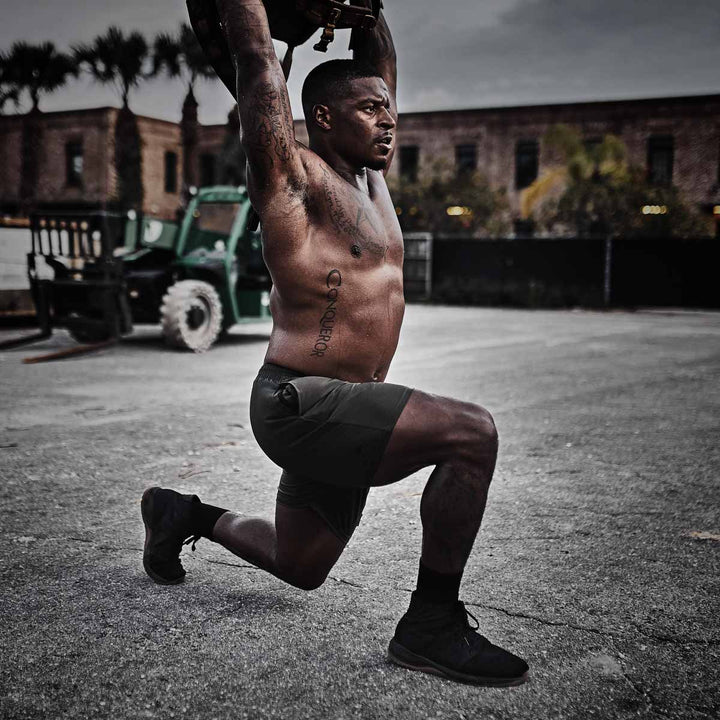 Wearing GORUCK's Ballistic Trainers in Black + Gum, a man executes an outdoor overhead lunge with weights on a cloudy day, demonstrating strength and balance.