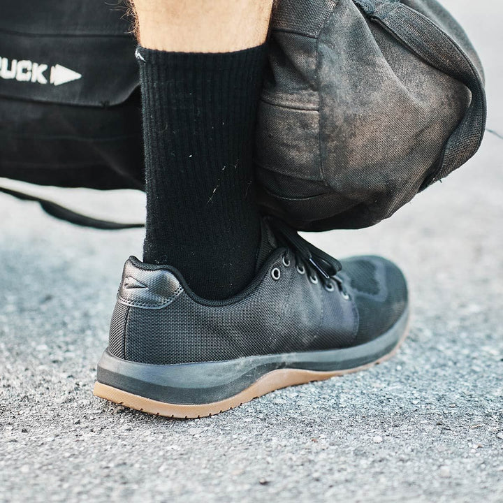 Close-up of a person wearing GORUCK's Ballistic Trainers in Black + Gum, showcasing their functional fitness footwear design, paired with socks. They're holding a black bag on a paved surface, possibly crafted from durable CORDURA® Ballistic Nylon.