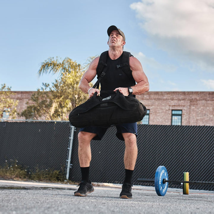 Wearing GORUCK's Ballistic Trainers in Black + Gum with a Black Reflective Spearhead, an athlete hoists a sandbag outdoors, sporting a cap and athletic gear. A blue weight plate rests on the ground nearby, while the durable CORDURA® Ballistic Nylon of their attire enhances the intensity of the workout session.