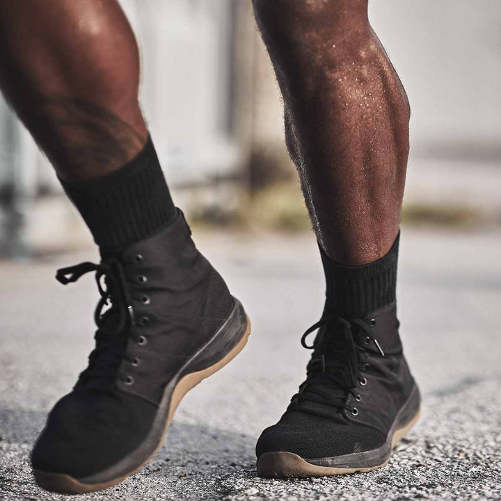 Close-up of a person wearing the GORUCK Ballistic Trainers - Mid Top in Black + Gum, made from durable CORDURA® Ballistic Nylon, walking on a gravel path. These functional fitness shoes offer 3X Support™ for comfort and stability.