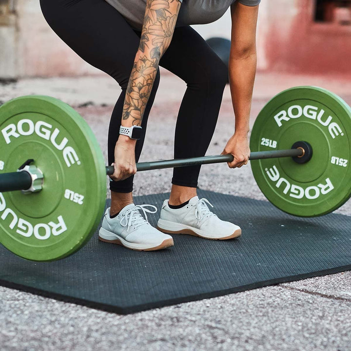 A person lifting a barbell with green weights on a black mat outdoors, showcasing the GORUCK Ballistic Trainers - Lunar Rock for maximum comfort and performance.