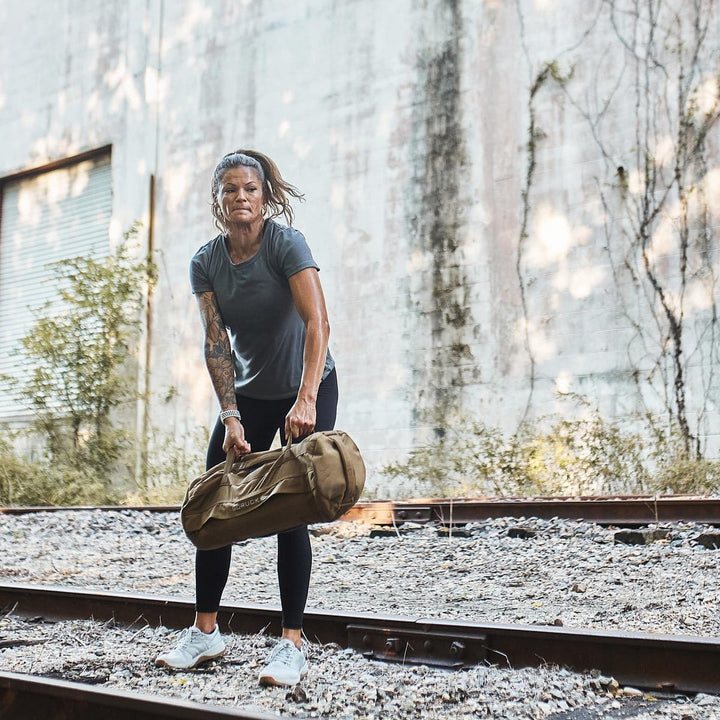 A person, demonstrating functional fitness, lifts a hefty bag on railway tracks with a worn wall in the background. Their GORUCK Ballistic Trainers in Lunar Rock offer durable support and reliability amid challenging conditions.