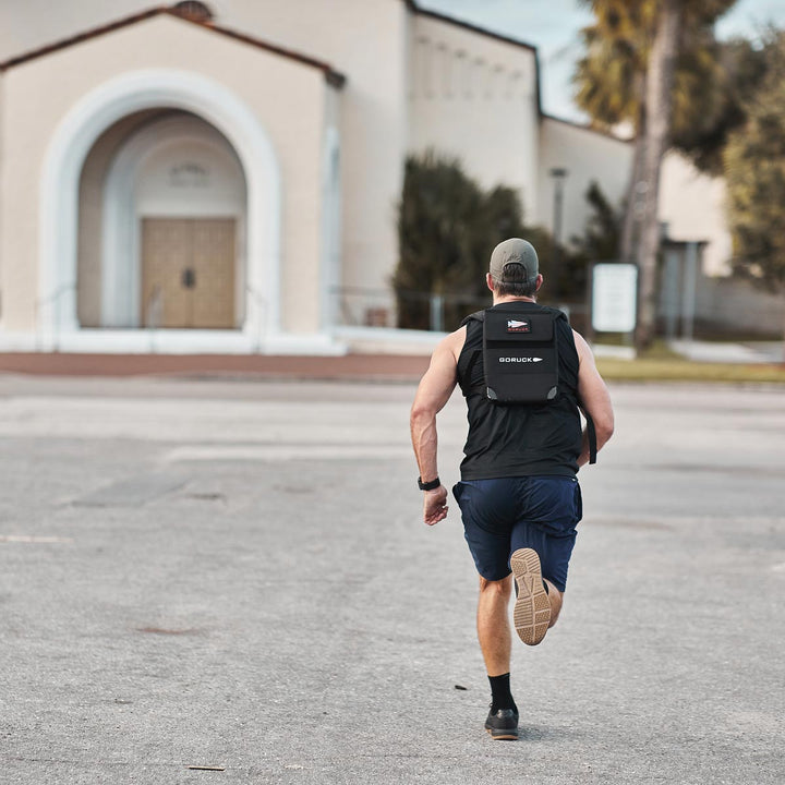 Laced in GORUCK's Ballistic Trainers - Black + Gum with the Black Reflective Spearhead and shouldering a backpack, a person dashes towards a white church building on a paved surface, their cap catching the breeze.