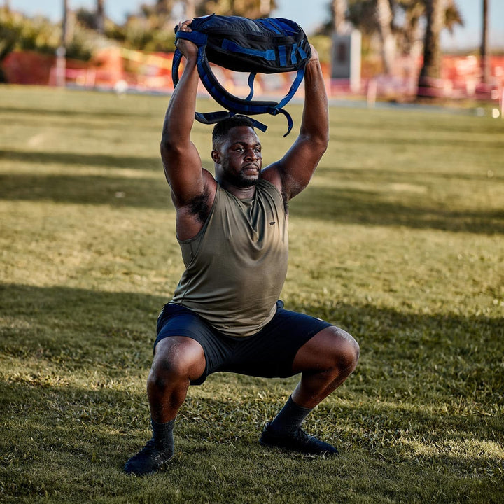 In a sunny park, a man in GORUCK's Ballistic Trainers - Blackout + Black does squats with a backpack held overhead. His workout shoes feature 3X Support™ for stability and comfort.