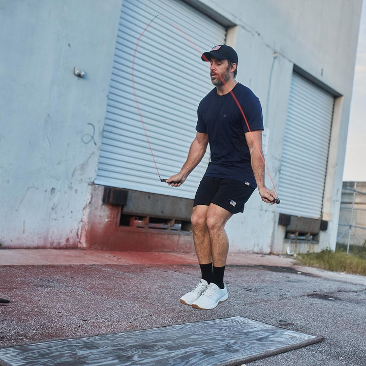 A man is jump roping outdoors near a building with large shutters, wearing the GORUCK Ballistic Trainers in Lunar Rock, crafted from CORDURA® Ballistic Nylon for maximum durability.