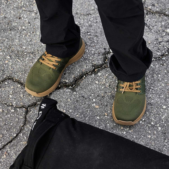 A person in dark pants wearing GORUCK's Ballistic Trainers - Green + Gum, specifically designed for functional fitness, stands on cracked pavement with a black bag nearby. Made from durable CORDURA® Ballistic Nylon, these trainers offer 3X Stability for those always on the move.