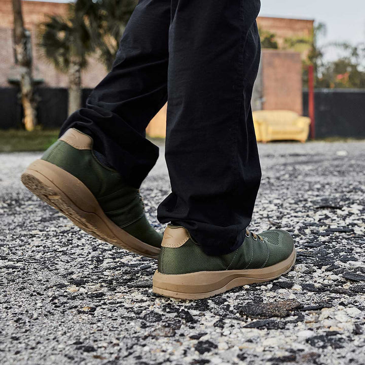 Close-up of someone walking on gravel wearing GORUCK Ballistic Trainers - Green + Gum paired with black pants.