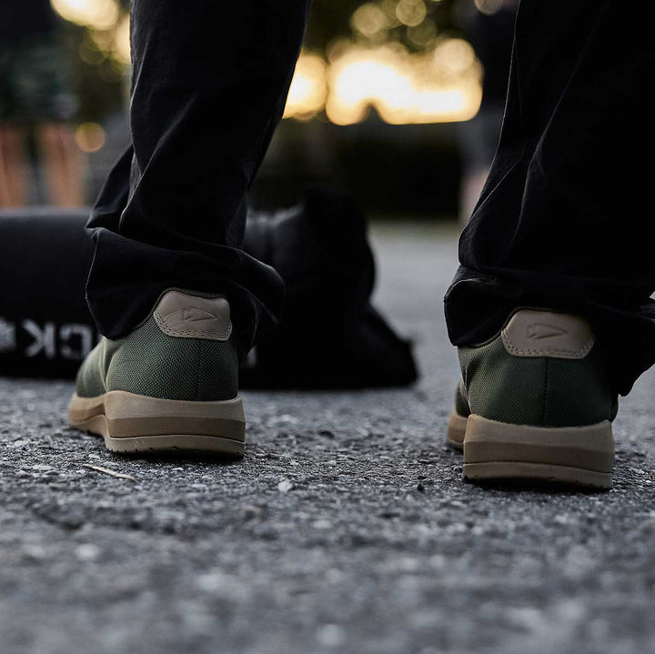 A person wearing GORUCK’s Ballistic Trainers - Green + Gum confidently strides along the gravel path, benefiting from the 3X Stability of their shoes. Nearby, a rugged duffel bag made from CORDURA® Ballistic Nylon awaits, ready for any adventure they embark upon.