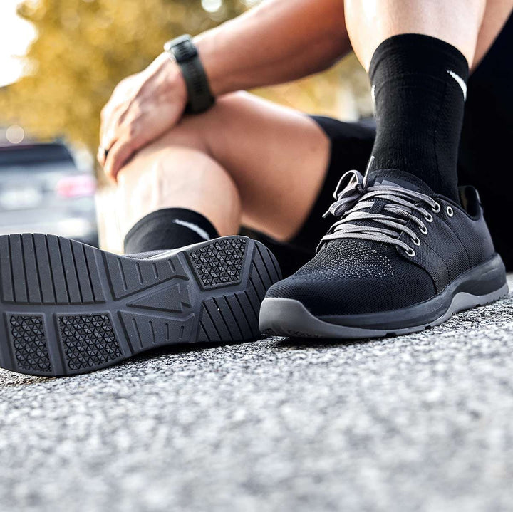 A person sits on the pavement wearing GORUCK Ballistic Trainers in black, black, and charcoal, crafted with CORDURA® Ballistic Nylon and paired with black socks, drawing attention to the durable shoe soles—perfect for individuals who prioritize functional fitness footwear.