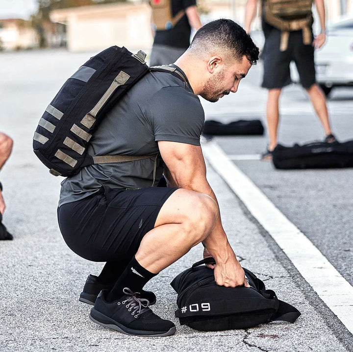 A man wearing GORUCK's Ballistic Trainers in Black, Black, and Charcoal is squatting outside, arranging bags crafted from CORDURA® Ballistic Nylon, with others visible in the background.