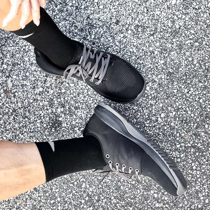 A person wearing GORUCK's Ballistic Trainers in Black + Black + Charcoal and black socks stands on a speckled pavement, showcasing functional fitness footwear designed with 3X Support™.