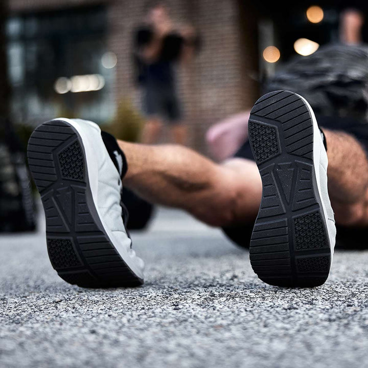 Close-up of a person lying on their back on asphalt, showcasing the soles of GORUCK's Ballistic Trainers in Lunar Rock + Charcoal, made from durable CORDURA Ballistic Nylon, ideal for functional fitness footwear.