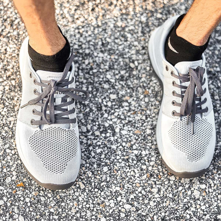 A person wearing GORUCK Ballistic Trainers in Lunar Rock and Charcoal, paired with black socks, stands on a textured pavement. The footwear is crafted from CORDURA Ballistic Nylon for functional fitness.