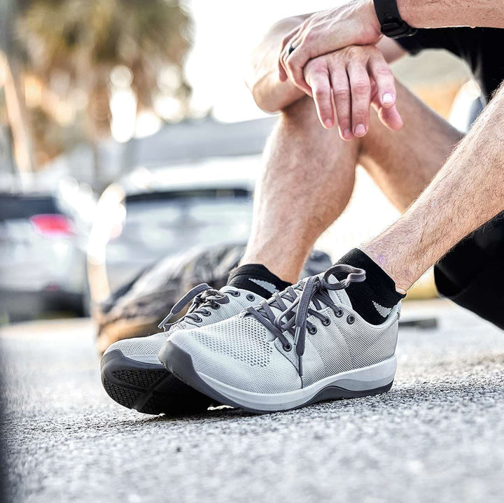 A close-up shows someone wearing GORUCK Ballistic Trainers in Lunar Rock and Charcoal, seated on a street with blurred cars behind them.