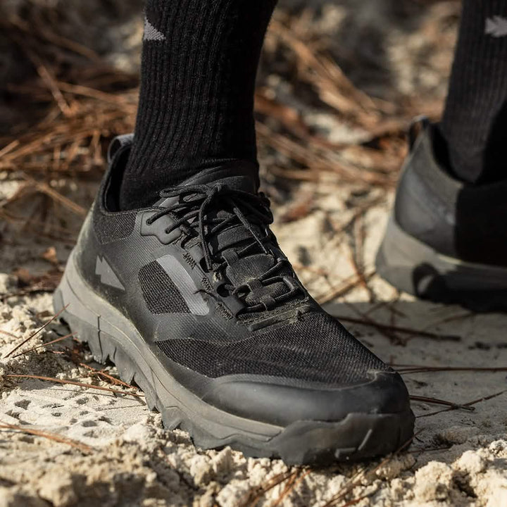 Close-up of a person wearing GORUCK's Mackall - Black + Forged Iron trail shoes with a triple compound outsole and black socks, standing on sandy ground scattered with pine needles.