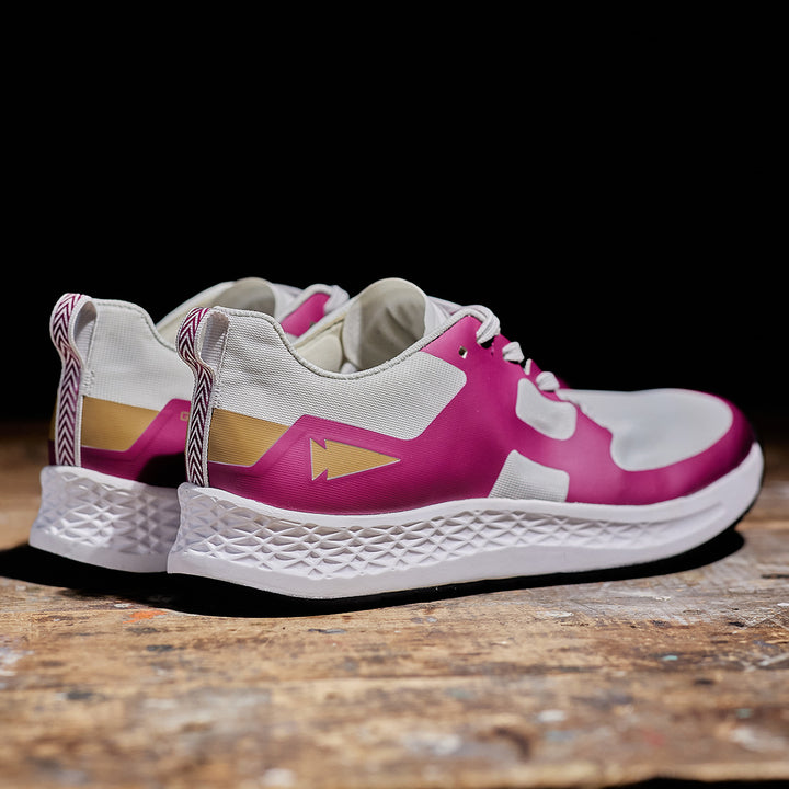 The Rough Runner - Baton Rouge running shoes by GORUCK, featuring a white and pink design with a road-to-trail outsole and textured soles, rest on a wooden surface. They are equipped with the innovative GRADIENT DENSITY™ EVA MIDSOLE for enhanced comfort and adaptability.