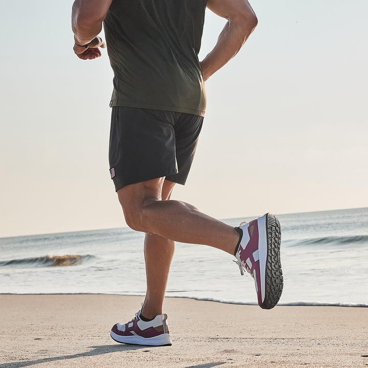 A person jogs along the beach, clad in a green shirt and black shorts, with waves crashing behind them. Their GORUCK Rough Runner - Baton Rouge shoes boast a road-to-trail outsole and GRADIENT DENSITY™ EVA MIDSOLE, ensuring comfort and support on the sandy terrain.