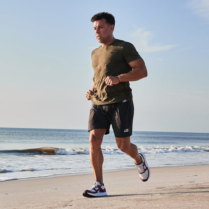 In the morning, a man jogs along the beach wearing a green shirt, black shorts, and GORUCK's Rough Runner - Baton Rouge shoes with a road-to-trail outsole and GRADIENT DENSITY™ EVA MIDSOLE for optimal support.