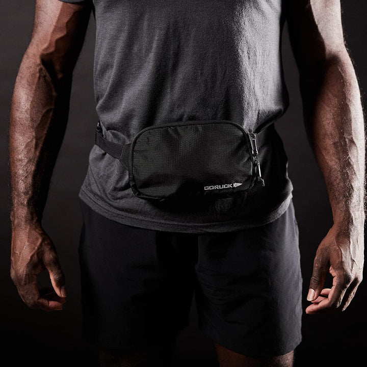 A person stands against a dark background wearing a gray shirt and black shorts, accessorized with the Belt Bag - Ripstop ROBIC® by GORUCK. This durable pack, made from sturdy Robic nylon, is enhanced with waterproof zippers for extra protection.