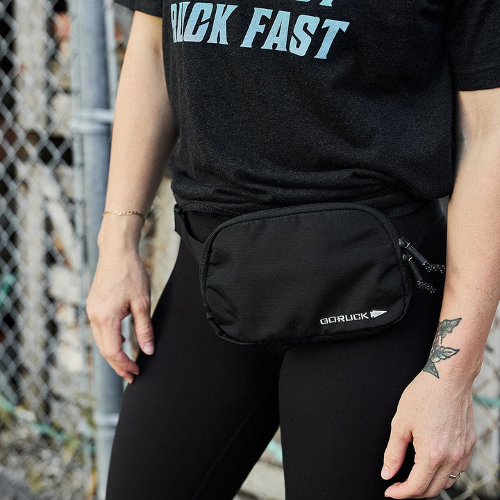 A person in a black T-shirt and leggings is outdoors near a chain-link fence, wearing a stylish GORUCK Belt Bag made from resilient Ripstop ROBIC® material. A small tattoo on their forearm adds a touch of personal flair.