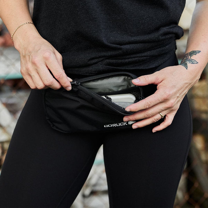 A person dressed in black leggings and a black shirt is holding a GORUCK Belt Bag - Ripstop ROBIC®. They are unzipping the waterproof zipper to reveal a white smartphone. The person has a tattoo on their left arm and wears a gold bracelet on their right wrist.