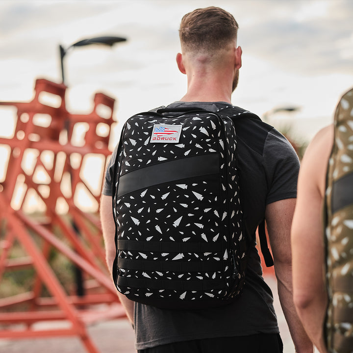 A person is wearing a black and white patterned GORUCK Rucker 4.0 rucksack, standing outdoors with red metal structures in the background.