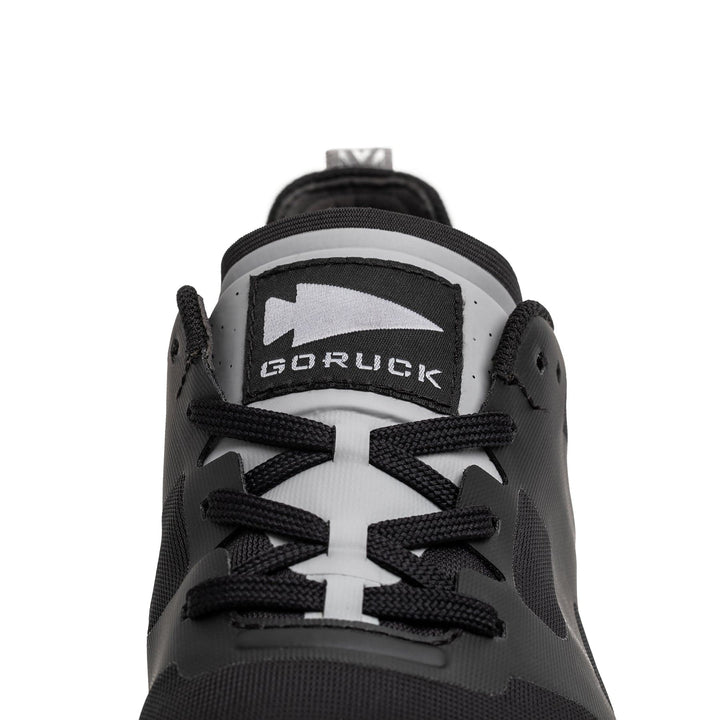 Close-up of a Men's Rough Runner sneaker by GORUCK in black and white featuring the logo on the tongue and black laces. This lightweight running shoe offers a sleek design with a gray stripe detail and includes a Gradient Density EVA Midsole for enhanced comfort.