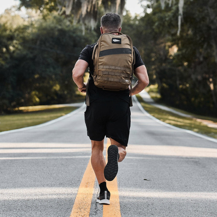 A person dressed in a black outfit with a brown backpack, wearing the Men's Rough Runner - Black + White by GORUCK, sprints down a two-lane road marked with a solid yellow line. Amidst the trees and greenery under clear skies, they embody the spirit of a Rough Runner in action.