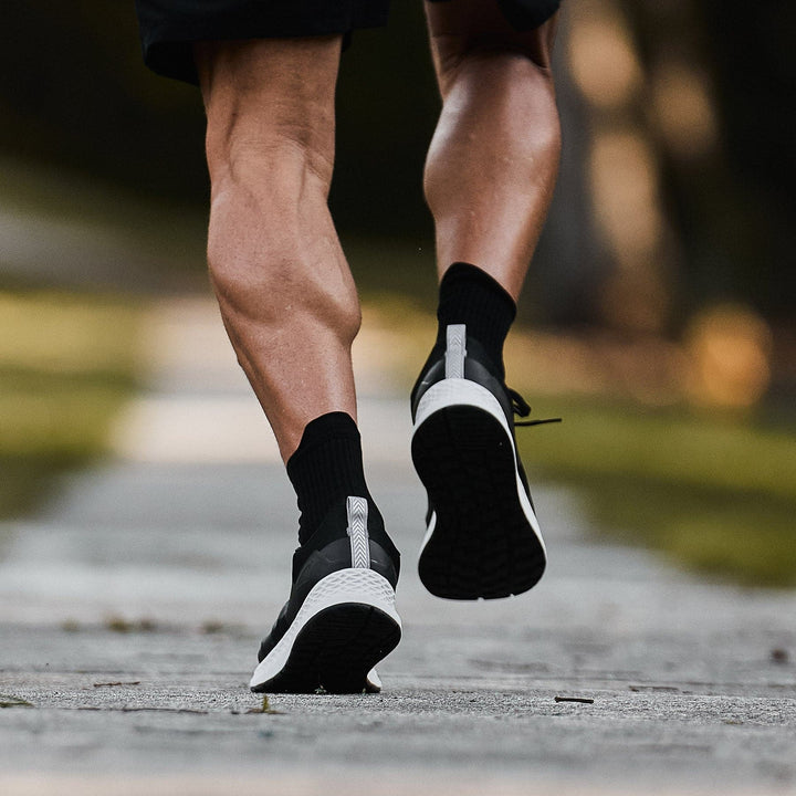 Close-up shot of a person sprinting on a paved path, with the focus on their legs and feet. They are wearing GORUCK's Men's Rough Runner shoes in black and white, featuring a Gradient Density EVA Midsole, along with black socks. The blurred background enhances the sense of dynamic movement similar to a Rough Runner challenge.