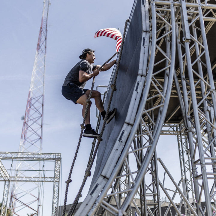 During the GORUCK Men's Rough Runner – Black + White outdoor obstacle course, a person climbs a steep black ramp with ropes. They reach for a red and white striped object at the top, surrounded by metal scaffolding under a clear blue sky, all while wearing lightweight running shoes for agility and comfort.