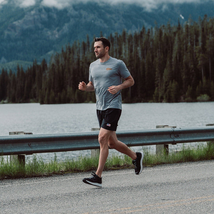 A man, famously called the "Rough Runner," jogs along a road beside a serene lake, surrounded by dense forest and misty mountains. He is outfitted in GORUCK's Men's Rough Runner - Black + White gear, which includes a gray t-shirt, black shorts, and running shoes equipped with a Gradient Density EVA Midsole. The overcast sky adds to the tranquil and natural setting.