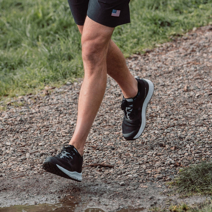 A person jogging on a gravel path in GORUCK Men's Rough Runner shoes in black and white, paired with shorts and carrying a small flag. Their legs are visible against a background of grass and a puddle, highlighting the superior comfort of the Gradient Density EVA Midsole, ideal for any Rough Runner.