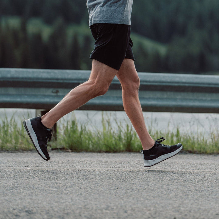 A person jogging on a paved road in Men's Rough Runner - Black + White by GORUCK, wearing black shorts and a gray shirt. The blurred greenery and metal guardrail in the background suggest a natural setting.