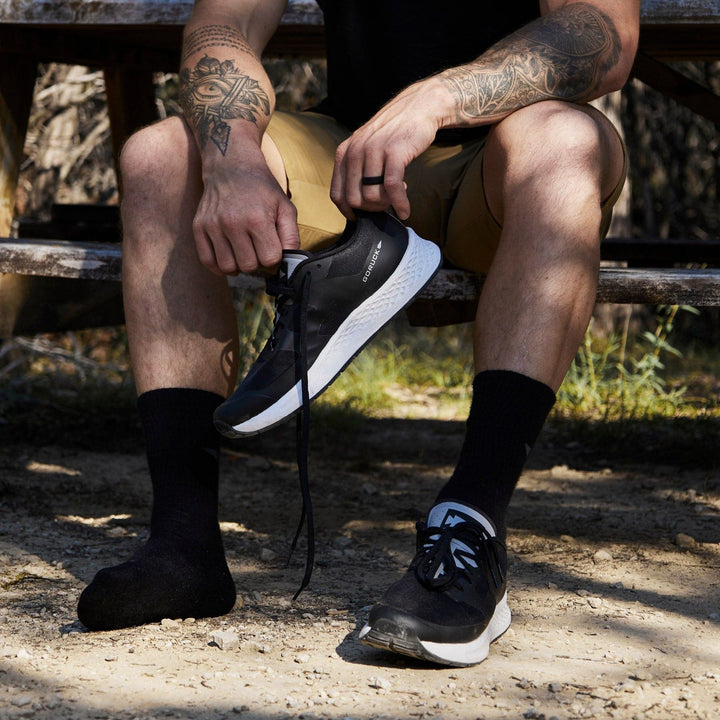 A person sitting on a wooden bench outdoors ties the laces of their GORUCK Men's Rough Runner shoes in black and white, perfectly paired with black socks and shorts. Sunlight filters through, highlighting the tattoos on their arms, while the Gradient Density EVA Midsole adds comfort for any adventurous run.