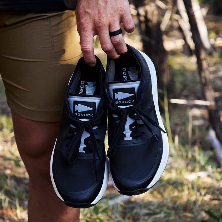 A person is holding a pair of Men's Rough Runner shoes in black and white by GORUCK, which include a Gradient Density EVA Midsole for comfort. They are dressed in tan shorts and a black ring. The outdoor setting, with grass and trees in the background, provides the perfect backdrop for these lightweight running shoes.