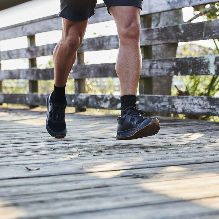 A person strolls across a wooden bridge in the Rough Runner - Black + Gum running shoes and shorts by GORUCK, with sunlight casting intricate shadows on the path.