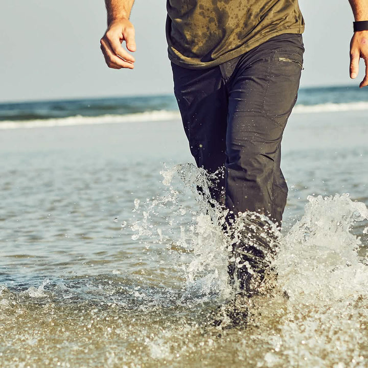 A person wearing GORUCK Men’s Challenge Pants - Lightweight ToughDry® and a damp olive green shirt strides through shallow ocean water, causing splashes, with the horizon visible in the background. The abrasion-resistant fabric keeps them comfortable while they wear a watch on their left wrist.