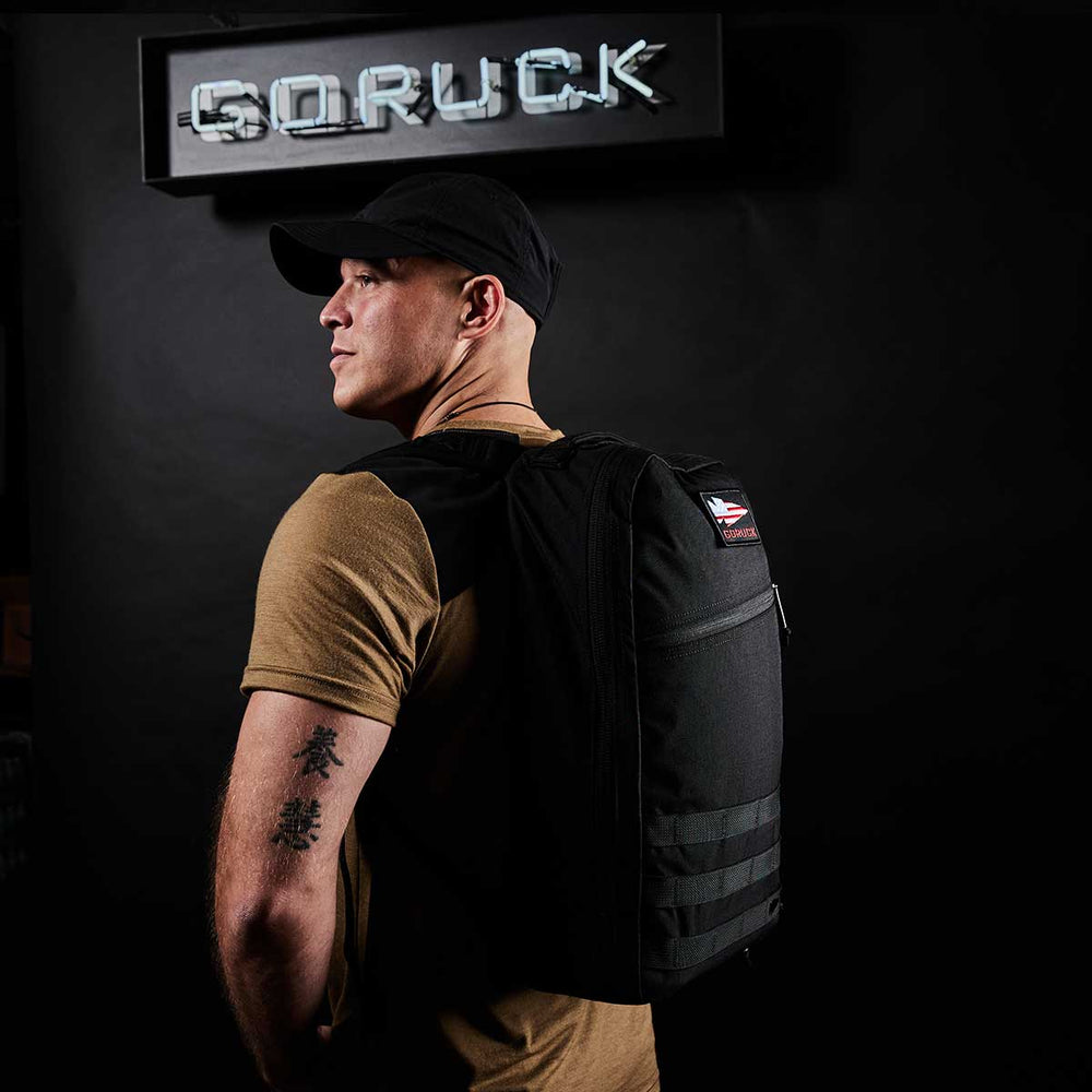 A person with a black cap and backpack stands in front of a "GORUCK" sign, showcasing the GORUCK Bullet Ruck Laptop - 16L, famed for its bombproof laptop compartment.