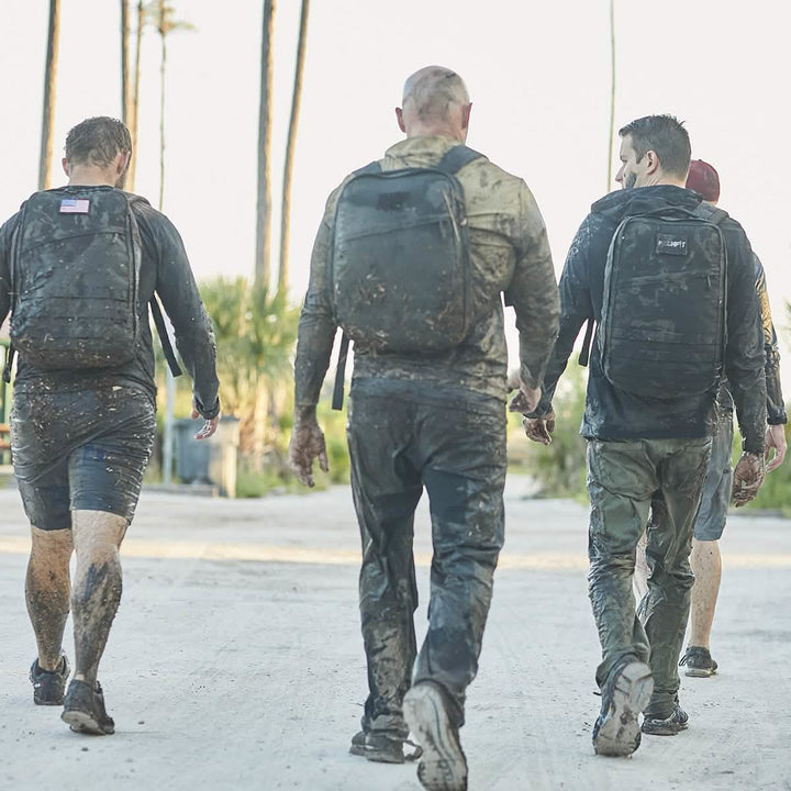 Four muddy individuals walk along a concrete path, wearing backpacks and casual outdoor attire that includes the lightweight Men’s Challenge Pants by GORUCK, featuring ToughDry® fabric for fast drying. Tall palm trees sway gently in the background under a clear sky.