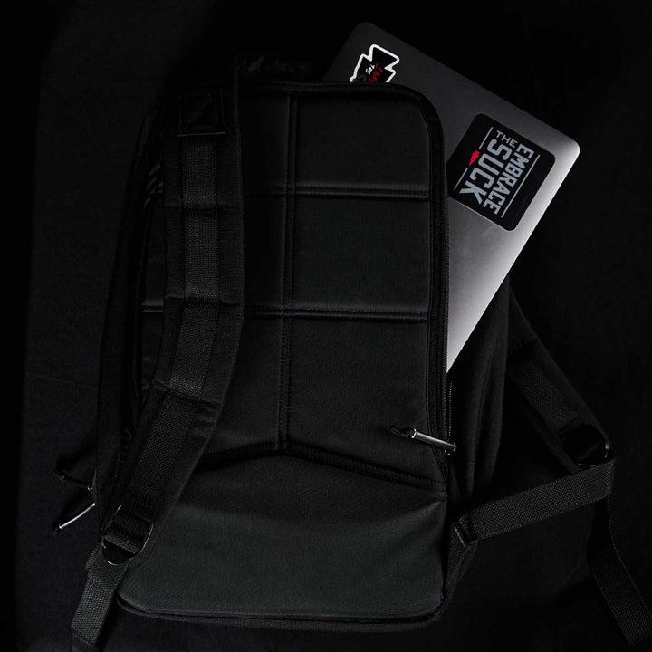 A Bullet Ruck Laptop - 16L by GORUCK, featuring a bombproof laptop compartment, reveals a sticker-adorned laptop peeking out of the black bag against a dark backdrop.