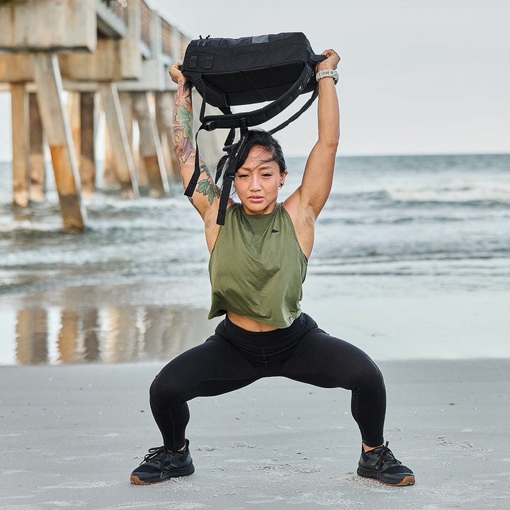 Dressed in athletic wear, a person executes a squat on a sandy beach while lifting a weighted Rucker 4.0 by GORUCK overhead. In the background, a pier stretches into the ocean as waves gently lap at the shore.