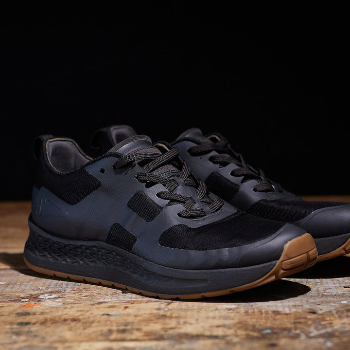 Resting on a wooden surface against a dark background are the Rough Runner - Black + Gum sneakers by GORUCK. These ultra-supportive running shoes feature a GRADIENT DENSITY™ EVA MIDSOLE, making them perfect for any adventure.