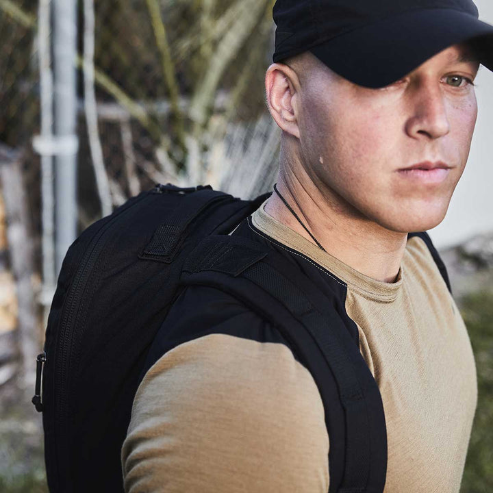 A person outdoors, wearing a black cap and carrying a GORUCK Bullet Ruck Laptop - 16L, which includes a bombproof laptop compartment, stands with a neutral expression.