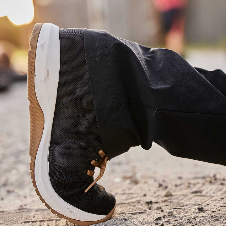 Close-up of a person's foot in GORUCK Ballistic Trainers in black and coyote, resting on a sandy surface outdoors, highlighting 3X Support for ultimate performance.