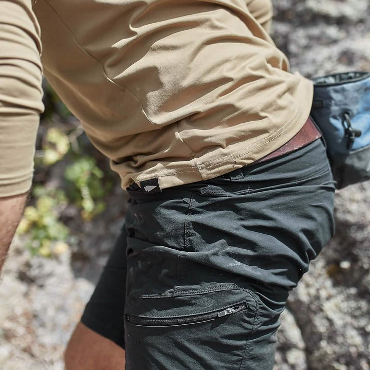 A person in a light brown long-sleeve shirt and GORUCK Men’s Challenge Shorts in black, made from Lightweight ToughDry® fabric with diamond gusset for enhanced comfort, is hiking outdoors. The focus is on their midsection and the shorts' cargo pockets, with a textured rock surface in the background. A gray bag is partially visible at their side.