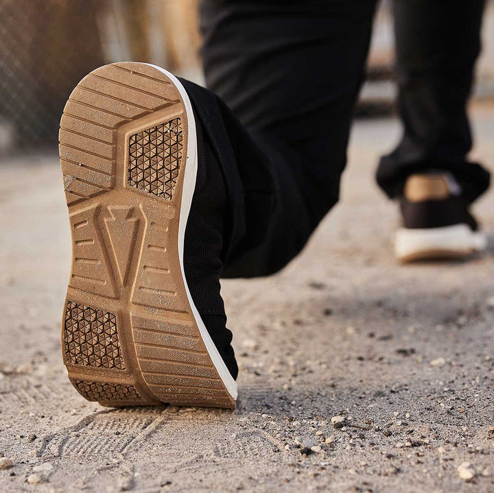 Close-up of a person wearing GORUCK Ballistic Trainers in Black, White, and Coyote as they walk on a sandy path, leaving distinctive footprints behind.