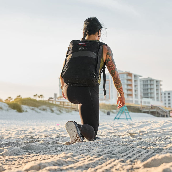 Kneeling on the sandy beach under a clear sky, with buildings rising in the background, a tattooed individual has a GORUCK Rucker 4.0 strapped to their back.