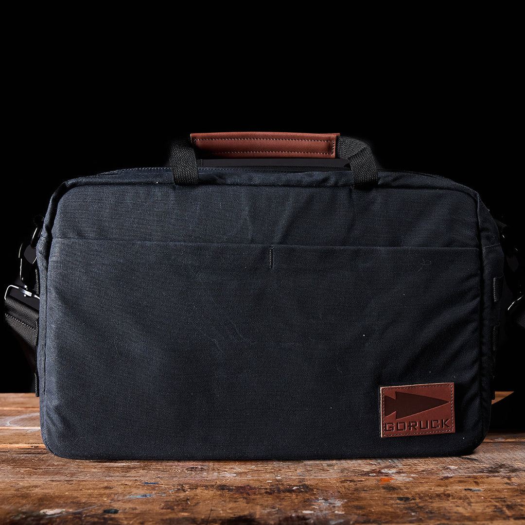 The Shoulder Bag - Waxed Canvas by GORUCK, crafted from black fabric with leather accents, includes a sturdy handle, shoulder strap, and front pocket. It is embellished with a brown leather patch featuring the GORUCK brand name in the bottom right corner. Placed on a wooden surface against a sleek black background, this bag is an essential accessory for any professional.